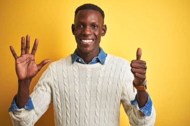 İzole sarı arka plan üzerinde kot gömlek ve beyaz kazak giyen Afrikalı Amerikalı adam gösteren ve parmak numarası altı ile işaret kadar emin ve mutlu gülümseyen.