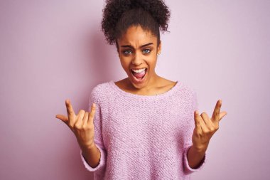Genç Afrikalı Amerikalı kadın el ile kaya sembolü yapıyor deli ifade ile izole pembe arka plan üzerinde ayakta kış kazak giymiş. Müzik yıldızı. Ağır müzik konsepti.