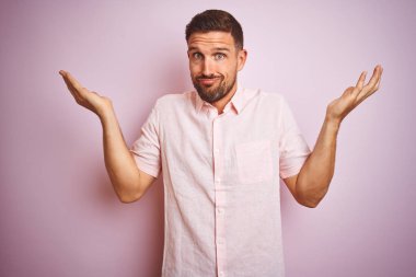 Pembe izole arka plan clueless ve kollar ve elleri ile karıştı ifade üzerine zarif yaz gömlek giyen genç yakışıklı adam kaldırdı. Şüphe kavramı.