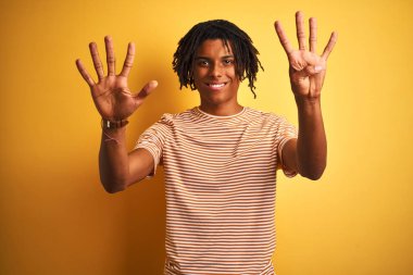 Dreadlocks ile izole sarı arka plan üzerinde duran çizgili t-shirt ayakta giyen ve emin ve mutlu gülümseyen iken parmak numarası dokuz ile işaret eden Afro adam.