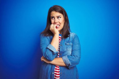 Genç güzel esmer kadın mavi izole arka plan üzerinde rahat mavi denim ceket giyen vurguladı ve ağız ısırma çivi eller ile gergin bakıyor. Anksiyete sorunu.