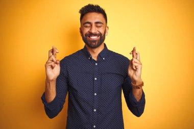 Genç yakışıklı Hint işadamı giyen gömlek üzerinde izole sarı arka planda işaret parmak karşı gülümsüyor umut ve gözler kapalı. Şans ve batıl fikir.