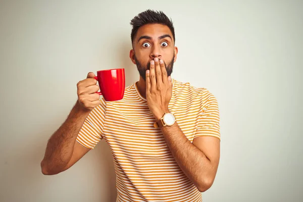 孤立した白い背景カバー口の上に立つ赤いコーヒーを飲む若いインド人男性は 間違い 恐怖の表現 沈黙の中で怖がって 秘密の概念でショックを受けた手で口を覆う — ストック写真