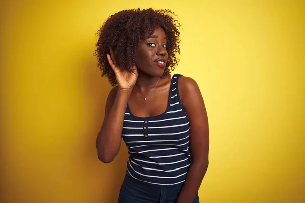 Young african afro woman wearing striped t-shirt over isolated yellow background smiling with hand over ear listening an hearing to rumor or gossip. Deafness concept.