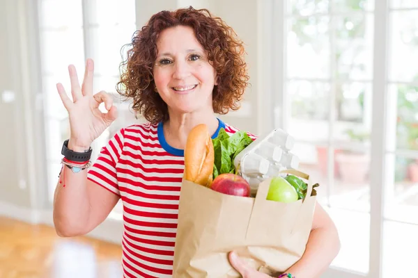 Senior woman holding paper bag full of fresh groceries from the supermarket doing ok sign with fingers, excellent symbol