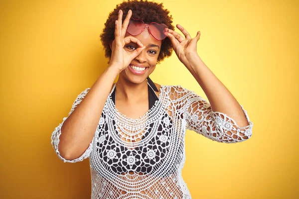 Young african american woman with afro hair wearing bikini and heart shaped sunglasses doing ok gesture with hand smiling, eye looking through fingers with happy face.