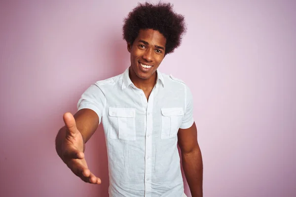 Young american man with afro hair wearing white shirt standing over isolated pink background smiling friendly offering handshake as greeting and welcoming. Successful business.