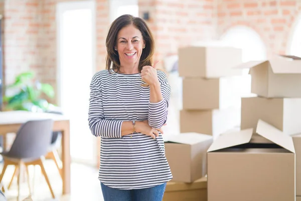 Middle age woman moving to a new house arround cardboard boxes smiling with happy face looking and pointing to the side with thumb up.
