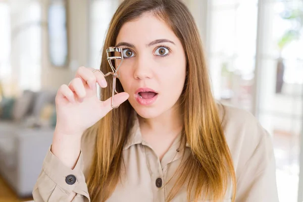 Beautiful young woman curling eyelashes using eyelash curler scared in shock with a surprise face, afraid and excited with fear expression
