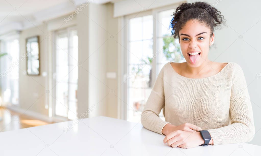 Beautiful young african american woman with afro hair sitting on table at home sticking tongue out happy with funny expression. Emotion concept.