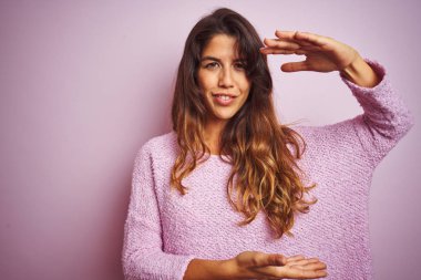 Genç güzel kadın pembe izole arka plan üzerinde duran kazak giyen elleri büyük ve büyük boy işareti gösteren gesturing, ölçü sembolü. Kameraya bakarak gülümsüyor. Ölçme kavramı.
