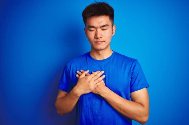 Genç Asyalı Çinli adam kapalı gözleri ve yüz minnettar jest ile göğsüne eller ile gülümseyen izole mavi arka plan üzerinde ayakta t-shirt giyiyor. Sağlık konsepti.