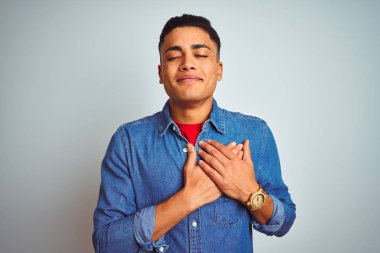 Genç Brezilyalı adam kapalı gözler ve yüz minnettar jest ile göğsüne eller ile gülümseyen izole beyaz arka plan üzerinde ayakta denim gömlek giyiyor. Sağlık konsepti.