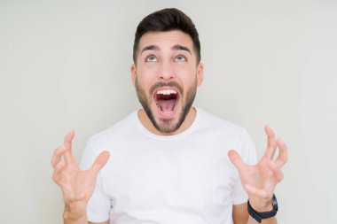 Üzerinde rahat beyaz t-shirt giyen genç yakışıklı adam arka plan deli izole ve deli bağırarak ve agresif ifade ve silah kaldırdı bağırıyor. Hayal kırıklığı kavramı.