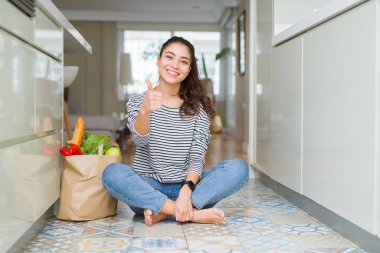 Genç kadın bir kağıt çanta taze bakkaliye dolu ile mutfak katında oturan mutlu thumbs el ile jest yapıyor. Başarı göstererek kameraya bakarak ifadeyi onaylama.