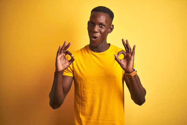 Young african american man wearing casual t-shirt standing over isolated yellow background looking surprised and shocked doing ok approval symbol with fingers. Crazy expression