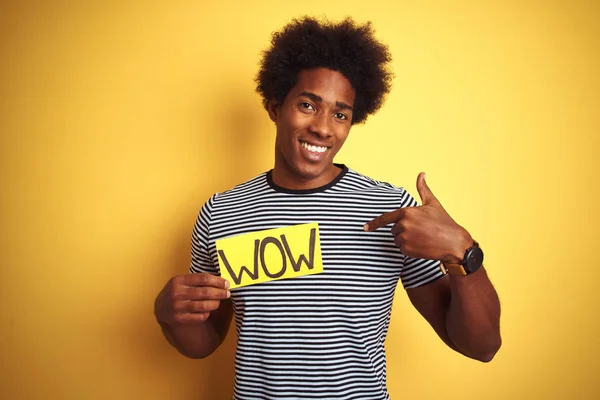 American man with afro hair holding wow banner standing over isolated yellow background with surprise face pointing finger to himself