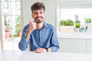 Beyaz masada oturan rahat gömlek giyen genç adam el ile jest kadar mutlu başparmak yapıyor. Başarı gösteren kameraya bakarak ifadeyi onaylama.