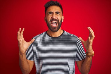 İzole kırmızı arka plan üzerinde lacivert çizgili t-shirt giyen genç yakışıklı Hint adam çılgın ve deli bağırma ve agresif ifade ve silah ile bağırdı kaldırdı. Hayal kırıklığı kavramı.