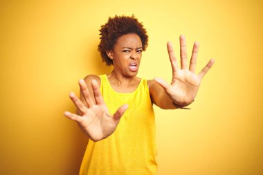 Beauitul Afrikalı Amerikan kadın izole sarı arka plan üzerinde yaz t-shirt giyiyor korkuyor ve korku ifadesi ile korkmuş el ile jest durdurmak, şok bağırıyor. Panik konsepti.