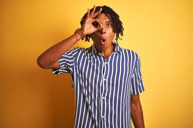 Kıvırcık sarı arka plan üzerinde rahat çizgili t-shirt giyen dreadlocks ile Afro adam sürpriz yüz, göz parmaklar ile bakarak şok ok jest yapıyor. İnanılmaz ifade.