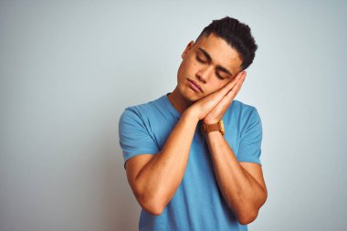 Genç Brezilyalı adam kapalı gözleri ile gülümseyen iken izole beyaz arka plan uyku yorgun hayal ve elleri ile poz üzerinde ayakta mavi t-shirt giyen.