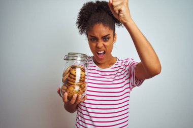 Genç Afrikalı Amerikan kadın izole beyaz arka plan üzerinde çerezleri bir kavanoz tutan rahatsız ve öfke ile bağırarak sinirli, deli ve yükseltilmiş el, öfke kavramı ile bağırarak