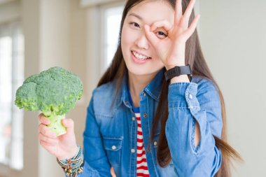 Güzel Asyalı kadın mutlu yüz gözleri parmaklarıyla görünümlü el ile ok işareti yapıyor gülümseyen Yeşil taze brokoli yeme