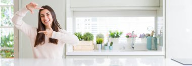 Güzel genç kadın evde beyaz masaya oturan geniş açı resim elle yapma çerçeve ve parmakları ile mutlu yüz gülümsemeye. Yaratıcılık ve Fotoğrafçılık kavramı.