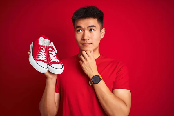 Young asian chinese man holding casual sneakers standing over isolated red background serious face thinking about question, very confused idea