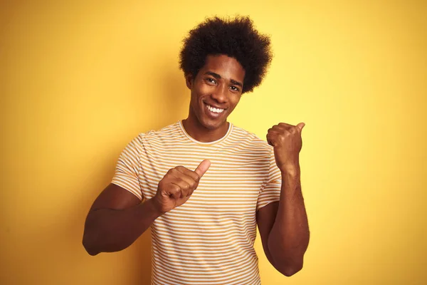 孤立した黄色の背景の上に立つストライプのTシャツを着たアメリカ人男性は 手と親指で後ろを指し 自信を持って微笑む — ストック写真