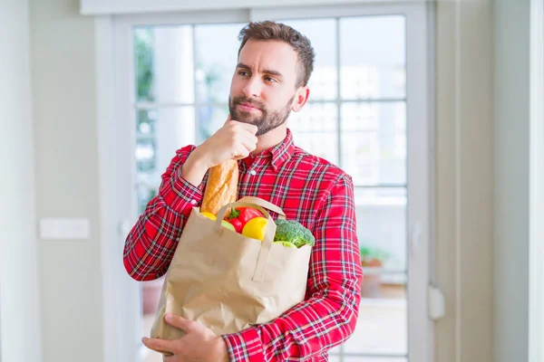 Handsome man holding groceries bag serious face thinking about question, very confused idea