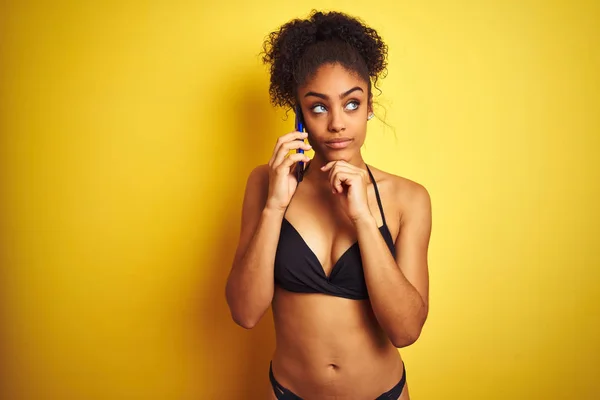 Afro american woman wearing bikini talking using smartphone over isolated yellow background serious face thinking about question, very confused idea