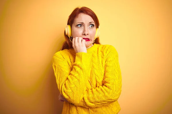 年轻的红发女郎在黄色孤立的背景上用耳机听音乐 双手咬着指甲 看起来紧张和紧张 焦虑问题 — 图库照片