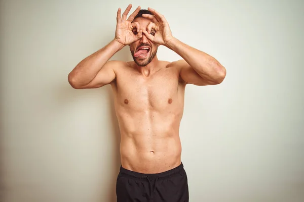 Young handsome shirtless man over isolated background doing ok gesture like binoculars sticking tongue out, eyes looking through fingers. Crazy expression.