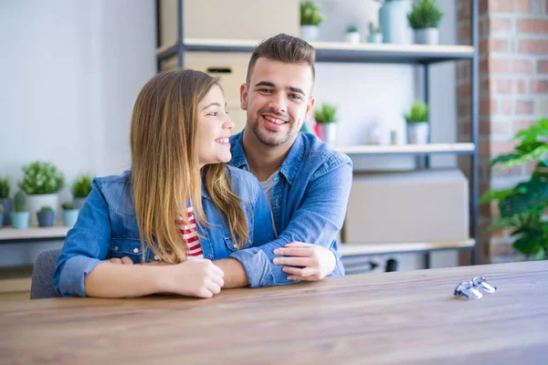 Young beautiful couple sitting on the table at home, hugging in love very happy for moving to new home with cardboard boxes behind them