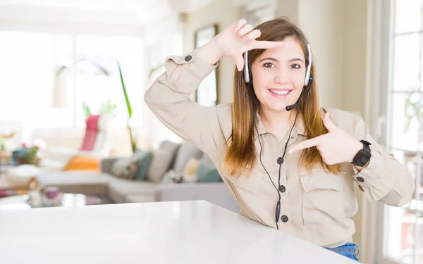 Beautiful young operator woman wearing headset at the office smiling making frame with hands and fingers with happy face. Creativity and photography concept.