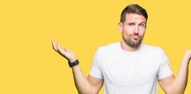 Yakışıklı adam rahat beyaz t-shirt clueless ve şaşkın bir ifadeyle kollar ve eller kaldırdı giyiyor. Şüphe kavramı.