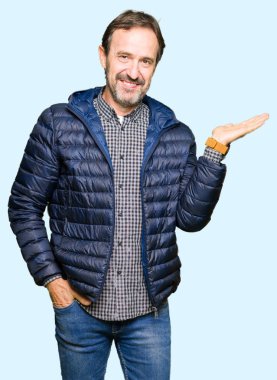 Orta yaş yakışıklı adam kış ceket giyen neşeli sunan gülümseyen ve el kamera bakarak avuç ile işaret.