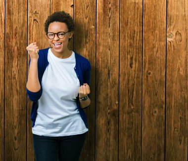 Gülümseyen ve başarı için çığlık izole arka plan çok mutlu ve heyecanlı kaldırdı, silah ile kazanan jest yapıyor üzerinde gözlük takan genç güzel Afrikalı-Amerikalı kadın. Kutlama kavramı.