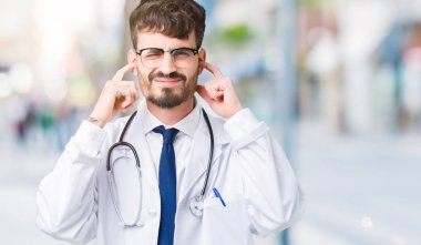 Genç doktor adam yüksek sesle müzik gürültü için rahatsız ifade ile parmaklar ile izole arka plan üzerinde hastane ceket giyiyor. Sağır kavramı.