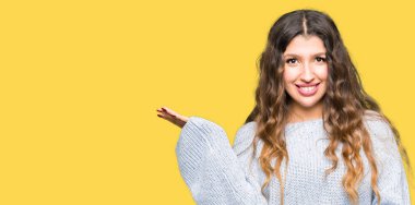 Neşeli sunulması ve kameraya bakarak avucunun ile işaret gülümseyen kış kazak giyen genç güzel kadın.