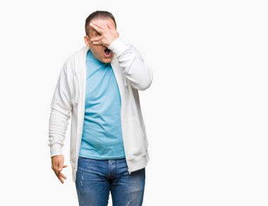Mahcup ifadeyle parmaklarımın arasından görünen yüz ve gözler el ile kapsayan şok izole arka plan bakmak üzerinde sweatshirt giyen orta yaş Arap adam.