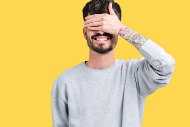 Genç yakışıklı adam gülümsüyor ve gözleri için sürpriz kapsayan yüz el ile gülüyor izole arka plan üzerinde kazak giyiyor. Kör kavramı.