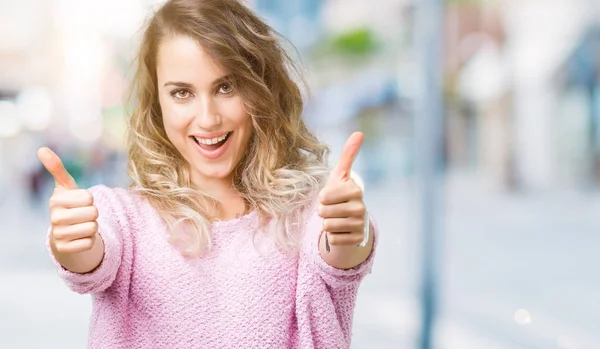 Beautiful young blonde woman over isolated background approving doing positive gesture with hand, thumbs up smiling and happy for success. Looking at the camera, winner gesture.