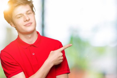 İzole arka planda kırmızı tişört giyen genç yakışıklı adam reklam, ciddi ve sakin yüz gösteren yan el parmağı ile Işaret