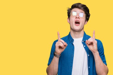 Yakışıklı delikanlı hayrete ve yukarıya bakıyor ve parmakları ile işaret ve silah kaldırdı sürpriz izole arka plan üzerinde gözlük.