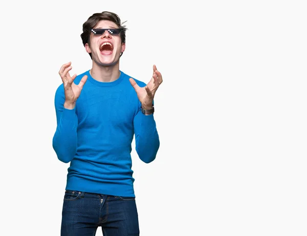 Young man wearing funny thug life glasses over isolated background crazy and mad shouting and yelling with aggressive expression and arms raised. Frustration concept.