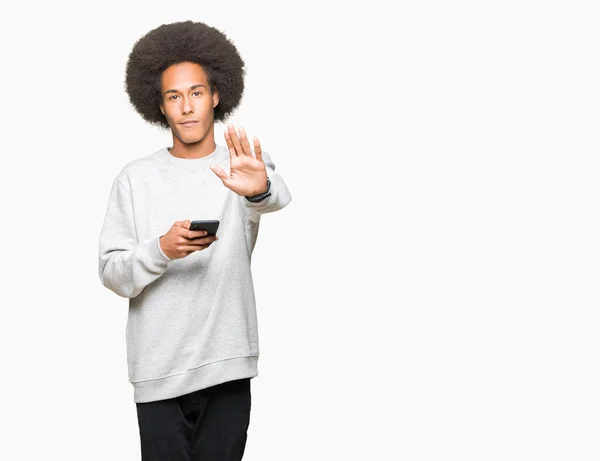 非洲青年美国人与非洲头发使用智能手机与开放的手做停止标志与严肃和自信的表达 防御姿态 — 图库照片