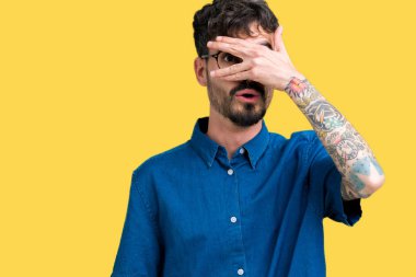 Mahcup ifadeyle parmaklarımın arasından seyir yüz ve gözler el ile kapsayan şok bakmak izole arka plan üzerinde gözlük takan genç yakışıklı adam.